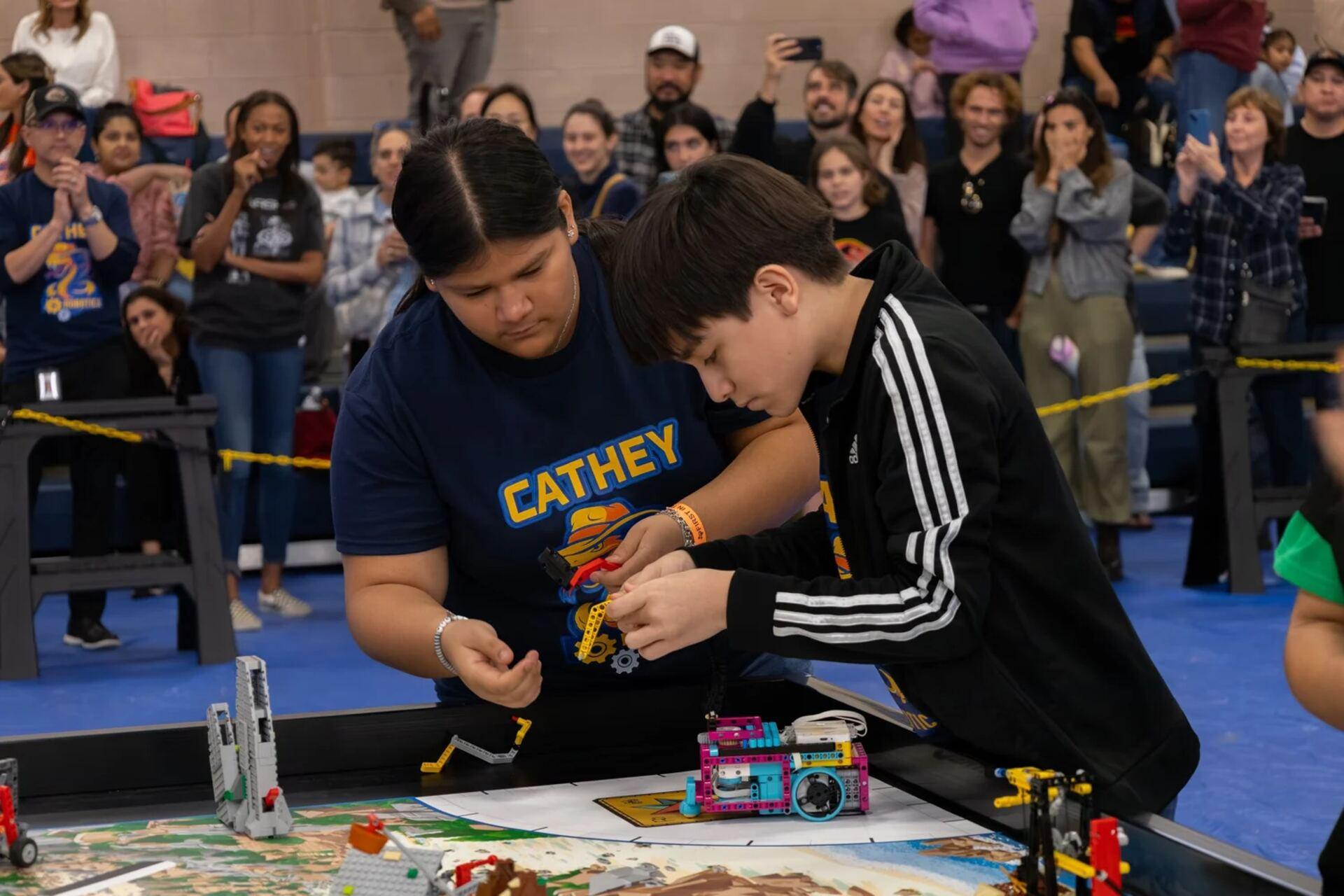 Two students reaching over a FIRST LEGO League table to set up their LEGO robot on the printed mission mat